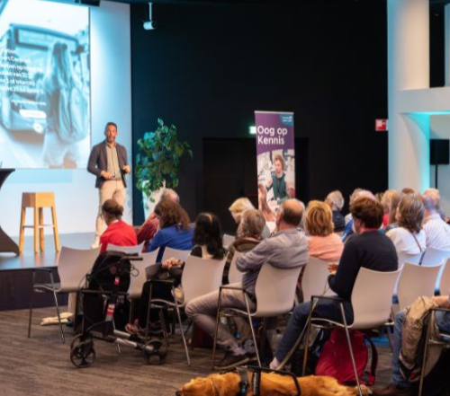 Bezoekers luisteren naar een spreker tijdens het kennisevent Oog op Kennis. De spreker staat op een podium voor een presentatie, terwijl deelnemers aandachtig in de zaal zitten. Op de achtergrond staat een banner van Oog op Kennis. Voorin ligt een hulphond rustig op de grond.