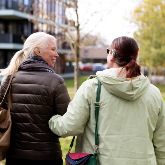 Twee personen lopen arm in arm over een voetpad langs een woongebouw. Op de achtergrond zijn bomen, gras en een rustige woonomgeving zichtbaar.
